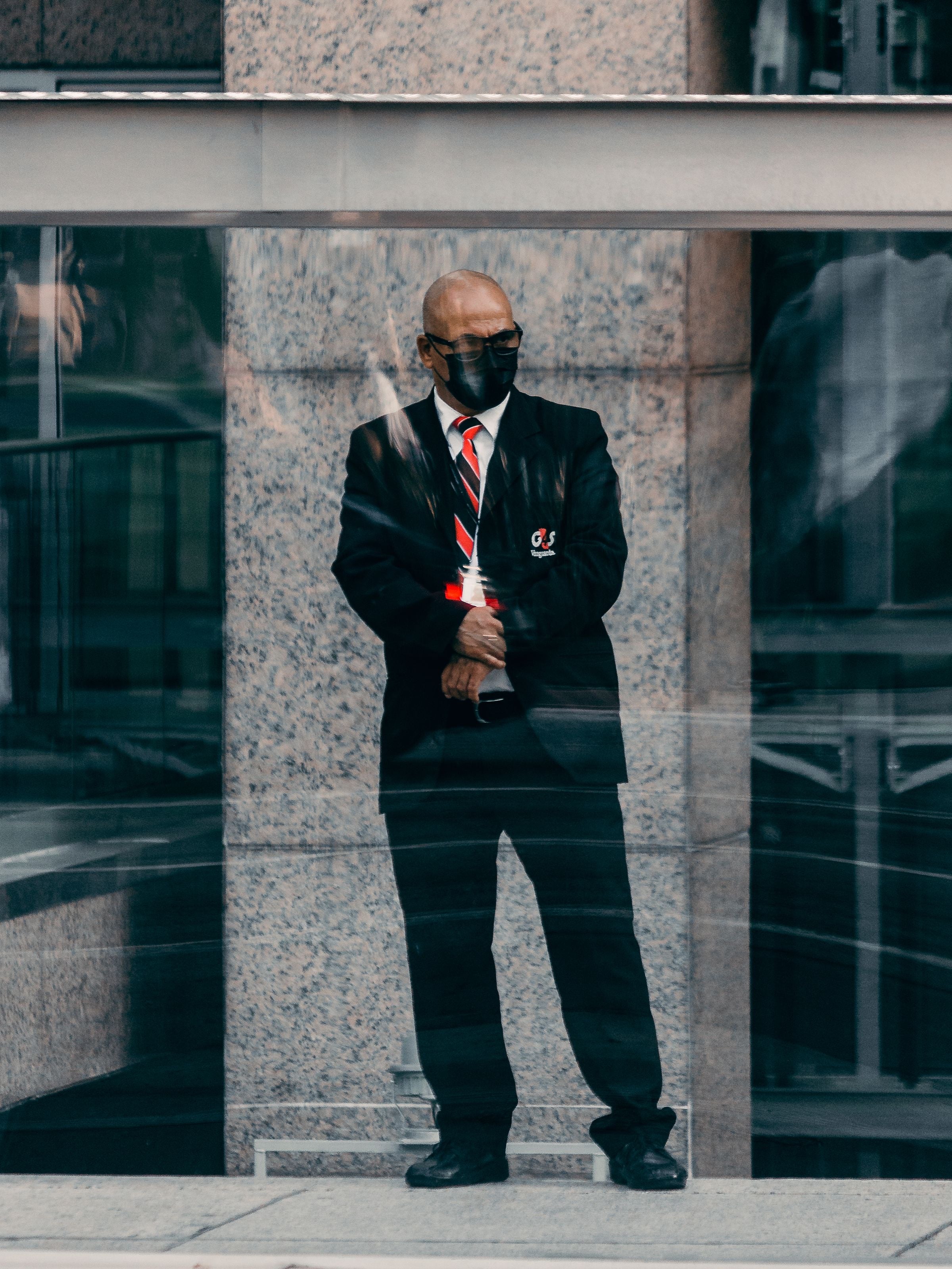 A security officer in a black suit and striped tie stands on duty at the glass entrance of an office building