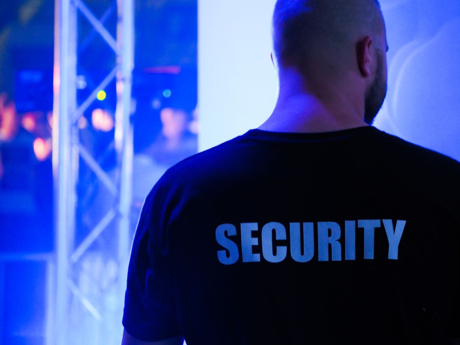 A door supervisor in a black T-shirt printed with SECURITY, watching a nightclub under blue lights