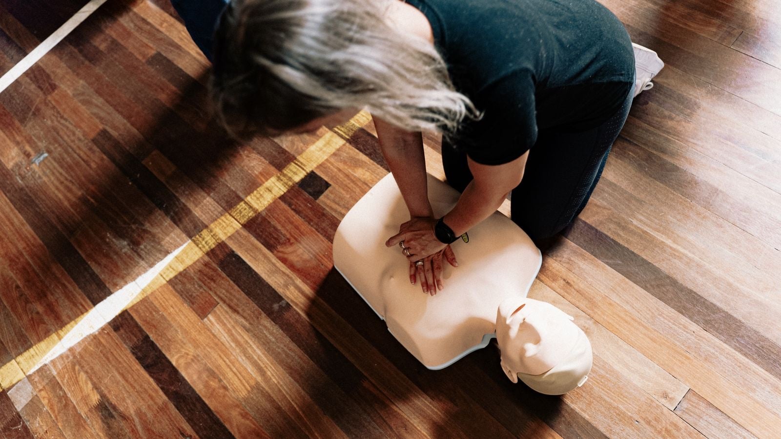 A trainee kneeling on a wooden floor practising chest compressions on a CPR mannequin