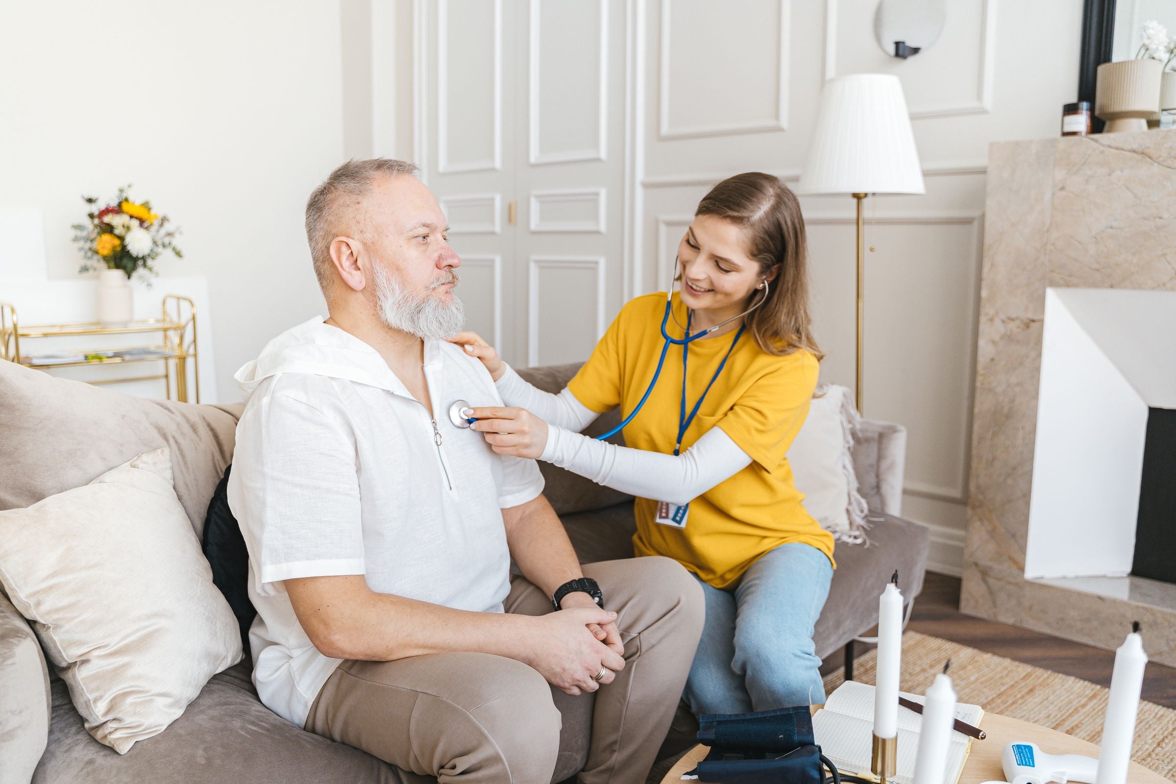 A care worker in a yellow top with an ID lanyard sitting beside an older man on his sofa at home