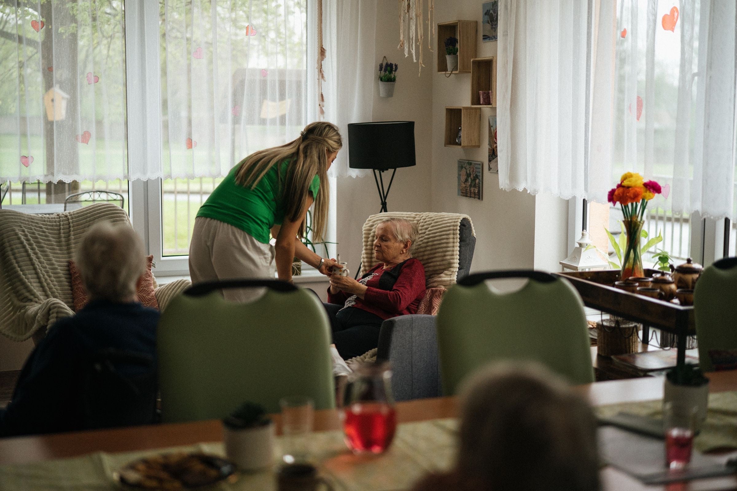 A care worker in a green top bending to talk with a resident in an armchair in a bright care home lounge