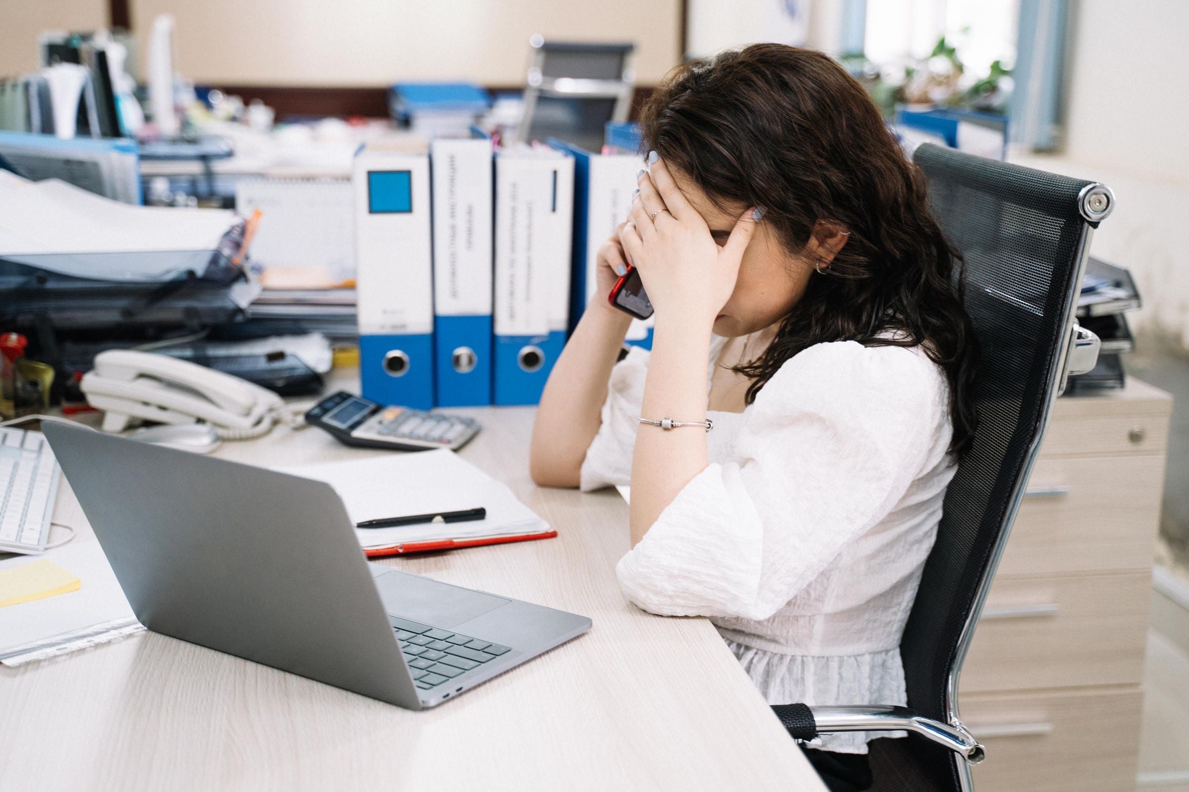 A coordinator at a busy office desk with her head in her hand during a phone call, folders and a laptop beside her